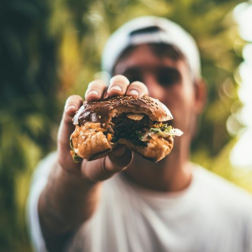 A man holds a juicy cheeseburger outdoors in daylight, highlighting its deliciousness and freshness.
